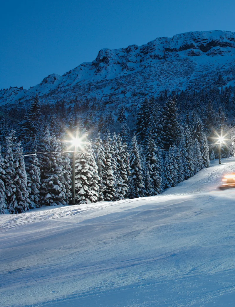 Blick über die beleuchtete Skipiste bei Nacht mit einer verschneiten Berglandschaft im Hintergrund.