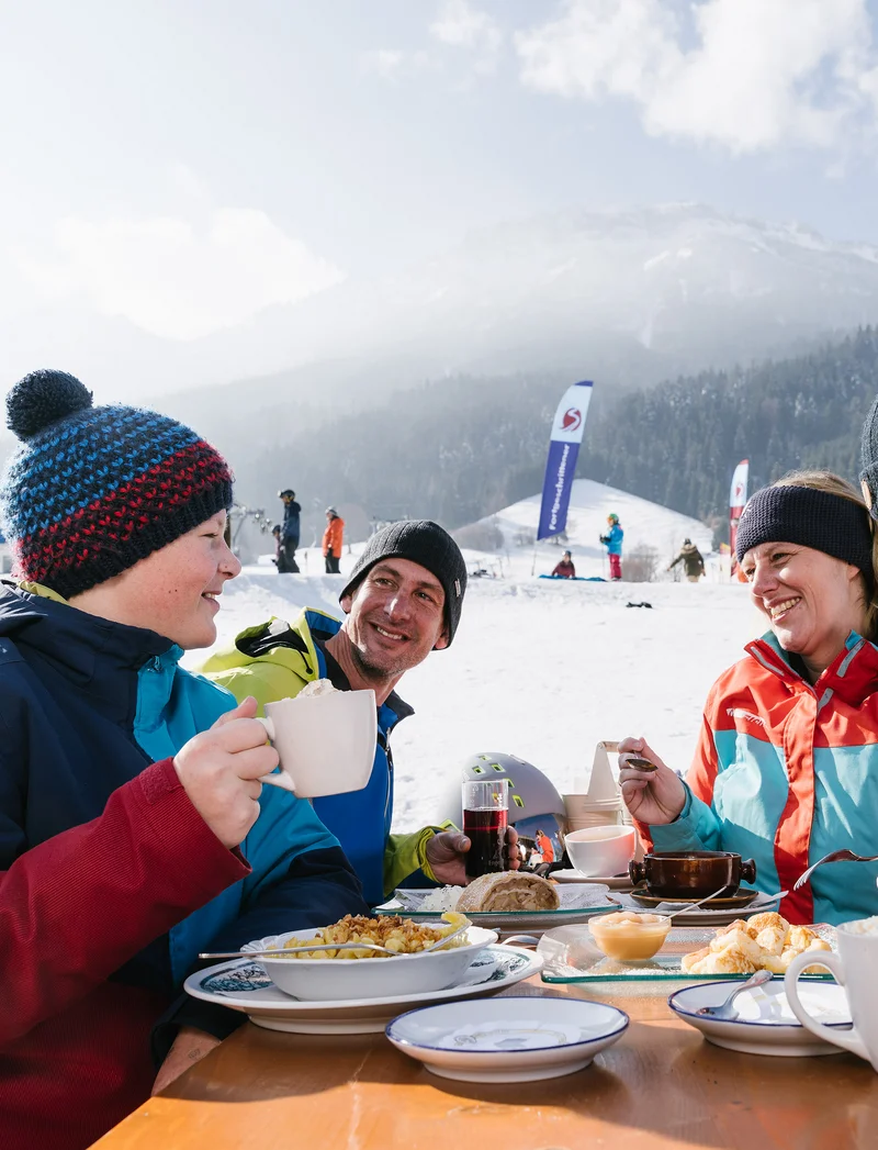 Eine Familie bei der Einkehr an der Skipiste, im Hintergrund eine verschneite Landschaft.