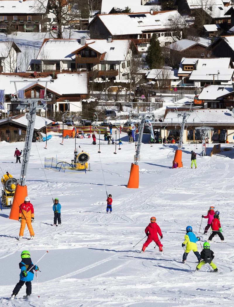 Kinder fahren die Skipiste hinunter, neben ihnen ist ein Tellerlift zu sehen.