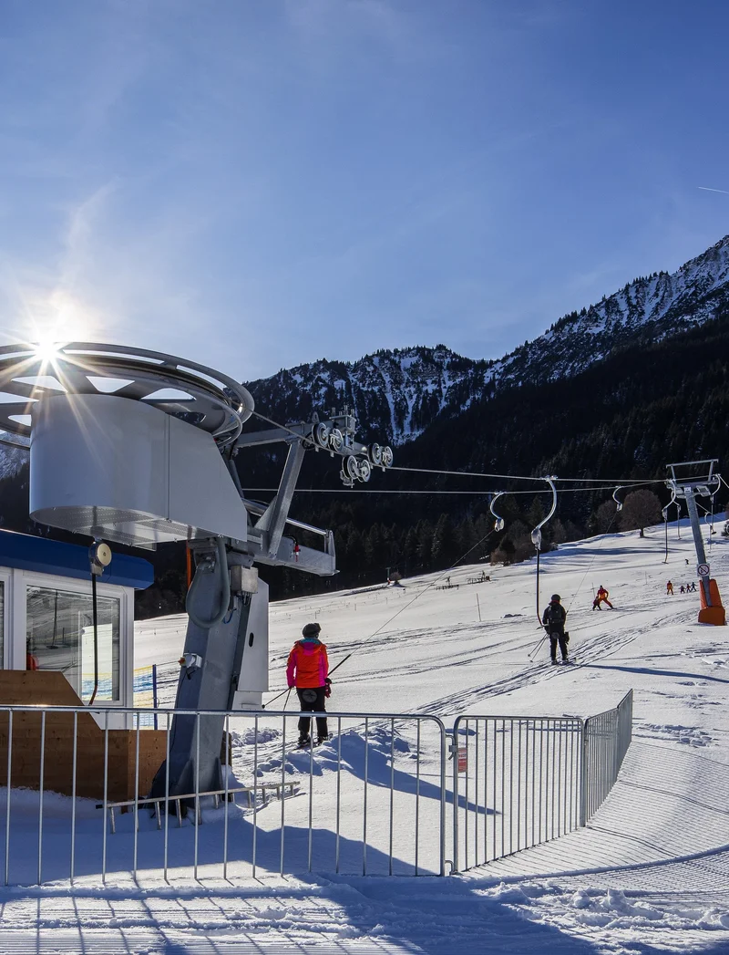 Skifahrer fahren mit einem Tellerlift, im Hintergrund ist eine weite Schneelandschaft zu sehen.